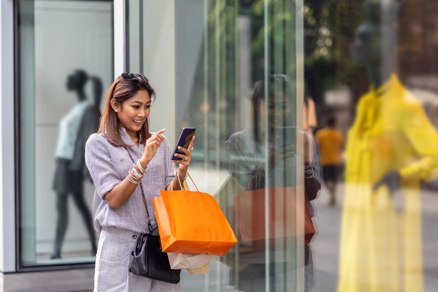 A woman with medium-length hair stands outside, smiling while looking at her smartphone. She is carrying several shopping bags, including an orange one. Dressed casually in a grey outfit and standing near a glass window that reflects her image, she wonders how to spend less money on future shopping trips. | LESSINVEST