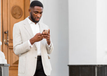 A man in a light gray blazer and white shirt is standing outside in front of a large building with white columns and a wooden door. He is looking down at his smartphone, possibly exploring the best investing apps. | LESSINVEST