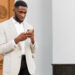 A man in a light gray blazer and white shirt is standing outside in front of a large building with white columns and a wooden door. He is looking down at his smartphone, possibly exploring the best investing apps. | LESSINVEST