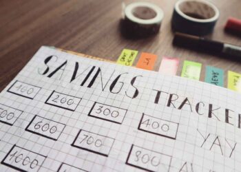 A budget savings tracker chart with hand-drawn grids and numbers is displayed on a table. The chart includes rectangular boxes for different amounts of money. Colorful sticky notes, two rolls of tape, and a red marker are seen in the background. | LESSINVEST
