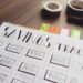 A budget savings tracker chart with hand-drawn grids and numbers is displayed on a table. The chart includes rectangular boxes for different amounts of money. Colorful sticky notes, two rolls of tape, and a red marker are seen in the background. | LESSINVEST