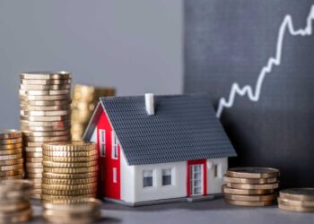 A small model house surrounded by stacks of gold coins illustrates the concept of investing in real estate. Behind them, a chalkboard displays an upward-trending graph, suggesting growth and financial gain from such investments. | LESSINVEST
