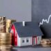 A small model house surrounded by stacks of gold coins illustrates the concept of investing in real estate. Behind them, a chalkboard displays an upward-trending graph, suggesting growth and financial gain from such investments. | LESSINVEST