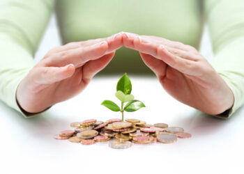 A pair of hands, positioned as if providing shelter, hover over a small green plant growing out of a pile of coins. The person, wearing a light green long-sleeve shirt, stands against a plain white background, emphasizing the concept of safe investments and financial growth. | LESSINVEST