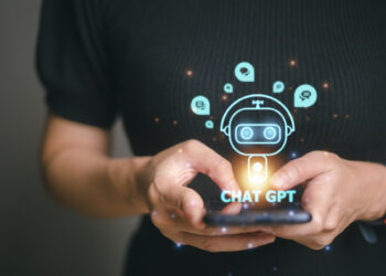 A person wearing a black shirt is holding a smartphone. A holographic image of a robotic face surrounded by social media icons appears above the phone, with "How to Invest in Chat GPT" written below the robot face. The person is tapping on the phone screen. | LESSINVEST