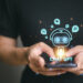 A person wearing a black shirt is holding a smartphone. A holographic image of a robotic face surrounded by social media icons appears above the phone, with "How to Invest in Chat GPT" written below the robot face. The person is tapping on the phone screen. | LESSINVEST