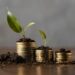 Three stacks of gold coins of varying heights are partly covered with soil, each boasting a green sprouting plant at the top. The blurred gray background emphasizes the coins and plants, symbolizing the best way to invest 10k for financial growth and sustainability. | LESSINVEST