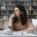 A woman with dark wavy hair wearing a beige blouse is sitting at a desk in a library, surrounded by open books and a laptop. Resting her chin on her hand, she stares pensively into the distance, embodying the essence of "invest in yourself." The background is filled with bookshelves. | LESSINVEST