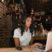A woman with long dark hair is focused on her laptop, researching how to make money online at a wooden table in a rustic, dimly lit café. She is wearing a white shirt and sitting next to a black mouse. The background includes brick walls, hanging lights, and various café items. | LESSINVEST