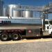A silver tanker truck from Waring Oil Company, LLC is parked at an oil depot, surrounded by large cylindrical storage tanks and industrial infrastructure. The truck has red and white safety placards indicating it's carrying hazardous materials—an everyday scene for those who choose to invest in oil. | LESSINVEST