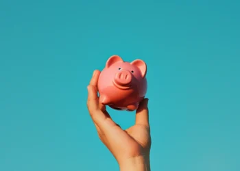 A hand is holding up a pink piggy bank against a clear, vibrant blue sky. With a cheerful expression, the piggy bank symbolizes the joy of savings and financial planning. | LESSINVEST