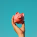 A hand is holding up a pink piggy bank against a clear, vibrant blue sky. With a cheerful expression, the piggy bank symbolizes the joy of savings and financial planning. | LESSINVEST