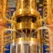 Close-up of a quantum computer's intricate interior, showcasing a complex network of gold-colored wiring and components. The central structure is surrounded by a variety of interconnected tubes and wires, forming an elaborate technological design that sparks curiosity about how to invest in AI. | LESSINVEST