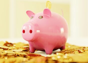 A pink piggy bank sits on an overflowing pile of gold coins, perfectly capturing the essence of save money. A coin is partially inserted into the slot on its back. The background is softly blurred in warm tones, emphasizing the piggy bank and coins in the foreground. | LESSINVEST