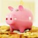 A pink piggy bank sits on an overflowing pile of gold coins, perfectly capturing the essence of save money. A coin is partially inserted into the slot on its back. The background is softly blurred in warm tones, emphasizing the piggy bank and coins in the foreground. | LESSINVEST