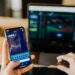 A person holding a smartphone displaying stock market graphs and numbers, with their other hand on a laptop keyboard. The laptop screen shows more charts and data, suggesting they are engaged in investing in ETFs or conducting financial analysis. | LESSINVEST