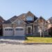 A two-story, suburban brick house with a gable roof, two-car garage, and a small front porch. The yard is bordered by trees, with a leafless tree slightly to the right. The house has a well-maintained lawn and is situated on a quiet residential street—ideal for investing in real estate. | LESSINVEST