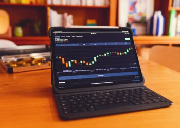 A tablet with a keyboard attachment is on a wooden desk. The screen displays a cryptocurrency price chart with various candlestick patterns, ideal for those investing in stocks. Two shelves with books and decorative items are blurred in the background. | LESSINVEST