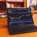 A tablet with a keyboard attachment is on a wooden desk. The screen displays a cryptocurrency price chart with various candlestick patterns, ideal for those investing in stocks. Two shelves with books and decorative items are blurred in the background. | LESSINVEST