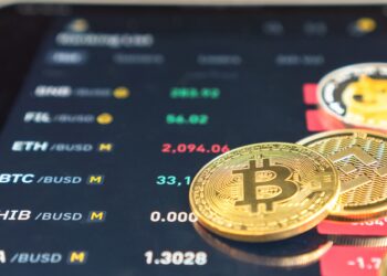 A close-up photograph shows a digital screen displaying various cryptocurrency prices in USD pairs, including BTC, ETH, ADA, and more. In front of the screen, several physical representations of cryptocurrencies, including a Bitcoin, illustrate the appeal of investing in crypto. | LESSINVEST