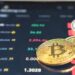 A close-up photograph shows a digital screen displaying various cryptocurrency prices in USD pairs, including BTC, ETH, ADA, and more. In front of the screen, several physical representations of cryptocurrencies, including a Bitcoin, illustrate the appeal of investing in crypto. | LESSINVEST
