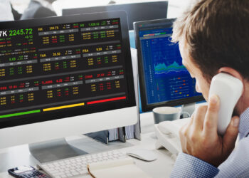 A man in business attire is on a phone call at his desk, facing dual computer monitors displaying stock market data from LessInvest.com Stocks to Invest In. Various indices, stocks to invest in, and prices are shown, with charts and numerical data featuring green and red indicators clearly visible on both screens. | LESSINVEST
