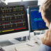 A man in business attire is on a phone call at his desk, facing dual computer monitors displaying stock market data from LessInvest.com Stocks to Invest In. Various indices, stocks to invest in, and prices are shown, with charts and numerical data featuring green and red indicators clearly visible on both screens. | LESSINVEST
