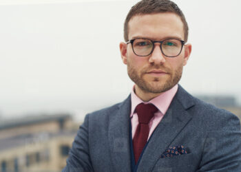 An entrepreneur with short hair and a beard is wearing glasses, a blue suit, a light pink shirt, and a dark red tie. He stands with his arms crossed, looking directly at the camera with a serious expression. The background is blurred, showcasing some buildings. | LESSINVEST