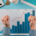 A person clenching their fists in excitement over a financial graph showing a steady upward trend from 2018 to 2021. The chart, coins, bills, a magnifying glass, and a notepad are placed on a blue desk, conveying financial growth and success through Invest Strategies by LessInvest.com Invest . | LESSINVEST