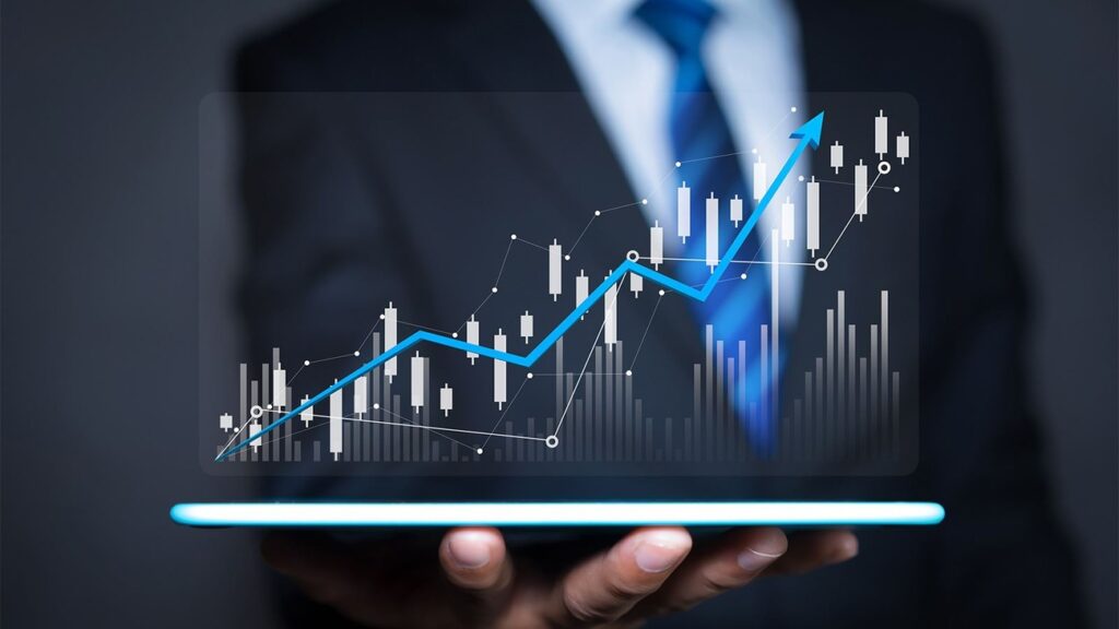A person in a suit holds a tablet displaying a glowing holographic graph with candlestick patterns and a rising trend line. The background is dark, emphasizing the financial chart, which suggests concepts of business growth, stock markets, and financial success. | LESSINVEST