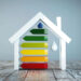 A house-shaped model illustrates how to make your home more energy efficient, featuring a vertical rating with colored bars from red at the bottom to green at the top. A water droplet symbol graces the right side, all set against a textured wooden surface. | LESSINVEST
