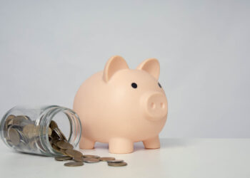 A pink piggy bank on a white surface is next to a tipped-over glass jar with scattered coins spilling out. The background is plain light gray. | LESSINVEST