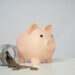 A pink piggy bank on a white surface is next to a tipped-over glass jar with scattered coins spilling out. The background is plain light gray. | LESSINVEST