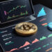 A smartphone displaying financial charts is docked, with a large gold Bitcoin coin resting on another screen showcasing crypto candlestick graphs and market data. The scene is set on a dark surface, exuding a tech-focused atmosphere. | LESSINVEST
