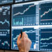 A person with glasses is analyzing multiple stock charts displayed on large screens. They are holding a pen and observing upward and downward trends on graphs, focusing on financial data and identifying stocks to invest in through detailed market analysis. | LESSINVEST
