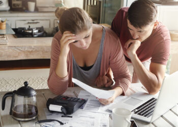A couple sits at a wooden table reviewing documents and discussing debt settlement on a laptop. The woman looks concerned, holding her head, while the man leans over to examine the papers. A coffee pot, calculator, and mug are on the table, with the kitchen visible in the background. | LESSINVEST