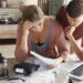 A couple sits at a wooden table reviewing documents and discussing debt settlement on a laptop. The woman looks concerned, holding her head, while the man leans over to examine the papers. A coffee pot, calculator, and mug are on the table, with the kitchen visible in the background. | LESSINVEST