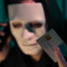 A person wearing a mask holds a credit card while talking on the phone, embodying the mysterious atmosphere often discussed in "Credit Card Fraud: Expert Tips from LessInvest.com." The card is held close to the camera, with numbers and details visible against a dimly lit background. | LESSINVEST