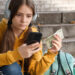 A millennial girl in a yellow hoodie sits on outdoor steps, wearing headphones and holding cash while checking her savings app on her phone. A green backpack is beside her. | LESSINVEST