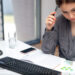 A woman sits at a desk, pen in hand, contemplating a colorful chart to make informed financial decisions. Surrounding her are essential tools: a keyboard, monitor, notebook, and smartphone. Nearby rests a glass of water, while the backdrop reveals sunlight filtering through window blinds. | LESSINVEST