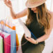 A woman in a hat and sleeveless dress sits outdoors, thoughtfully looking into her white purse. Surrounded by colorful shopping bags, she wears a watch on her left wrist, embracing the joy of smart style while choosing to spend less. | LESSINVEST