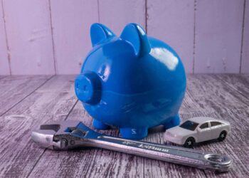 A blue piggy bank rests on a wooden surface alongside a miniature toy car and a metal wrench, embodying clever money-saving car hacks. The backdrop of a light wood-paneled wall completes this thoughtful arrangement. | LESSINVEST