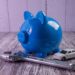 A blue piggy bank rests on a wooden surface alongside a miniature toy car and a metal wrench, embodying clever money-saving car hacks. The backdrop of a light wood-paneled wall completes this thoughtful arrangement. | LESSINVEST