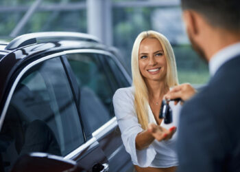 A woman with long blonde hair smiles as she receives car keys from a person in a suit, celebrating her new purchase. She stands next to a dark-colored vehicle, showcasing the joy of buying a car in a bright, modern indoor setting. | LESSINVEST