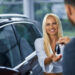 A woman with long blonde hair smiles as she receives car keys from a person in a suit, celebrating her new purchase. She stands next to a dark-colored vehicle, showcasing the joy of buying a car in a bright, modern indoor setting. | LESSINVEST