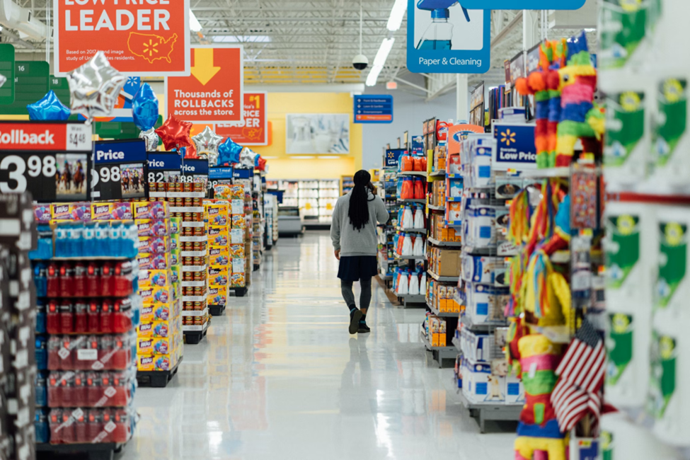 A person walks down an aisle in a brightly lit supermarket. Shelves are stocked with various products, including canned goods and cleaning supplies. Signs indicate "Low Price Leader" and "Thousands of Rollbacks. | LESSINVEST