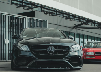 A black luxury sedan, its VIN visible on the windshield, is parked in front of a modern building. A red car is partially visible behind it. The setting exudes an urban flair with sleek, contemporary design. | LESSINVEST