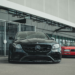 A black luxury sedan, its VIN visible on the windshield, is parked in front of a modern building. A red car is partially visible behind it. The setting exudes an urban flair with sleek, contemporary design. | LESSINVEST