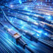 Close-up of glowing fiber optic cables emitting blue light. Several plugged connectors illustrate high-speed data transmission and internet connectivity, showcasing advanced internet infrastructure. The background is a blur of illuminated cables, suggesting fast network communication. | LESSINVEST