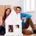 A smiling couple sits on the floor of a bright room surrounded by cardboard boxes, savoring the moment as they hold a large cutout of a white house. With wooden flooring and white walls, it feels like the best time to buy a home and start their new chapter. | LESSINVEST